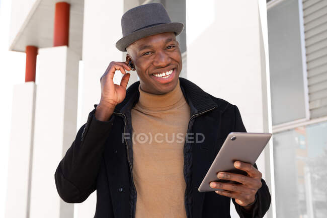 Homme afro-américain joyeux dans des vêtements élégants et écouteurs naviguant tablette moderne sur la rue ensoleillée et regardant la caméra avec le sourire — Photo de stock