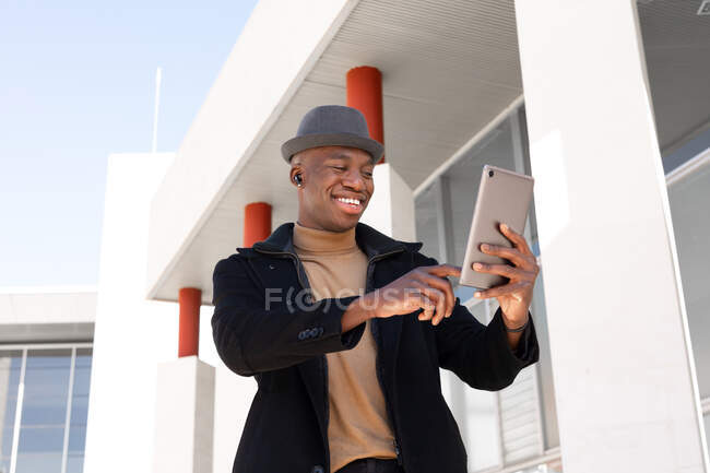 Allegro maschio afroamericano in abiti eleganti e auricolari navigando tablet moderno sulla strada soleggiata e guardando lo schermo con sorriso — Foto stock