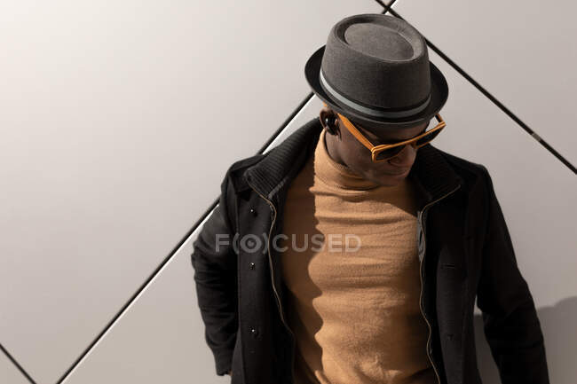 Hombre afroamericano confiado de moda en sombrero y gafas de sol de pie contra la pared gris y mirando hacia abajo - foto de stock