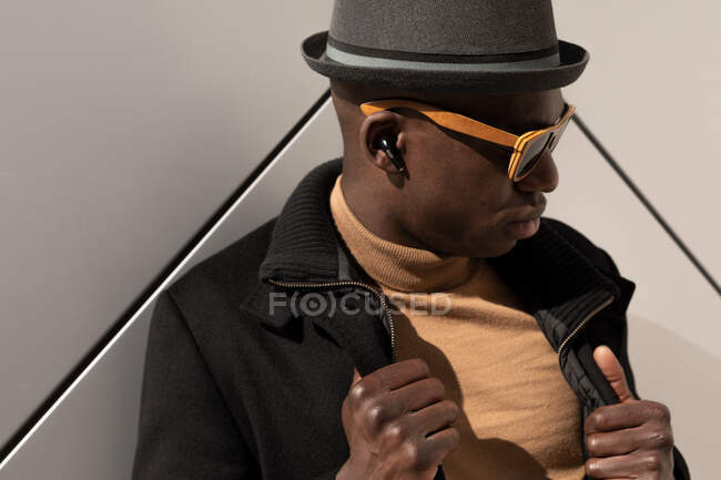 Hombre afroamericano confiado de moda en sombrero y gafas de sol de pie contra la pared gris y mirando hacia abajo - foto de stock