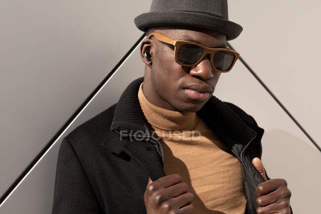 Trendy confident African American male in hat and sunglasses standing against gray wall and looking at camera — Stock Photo