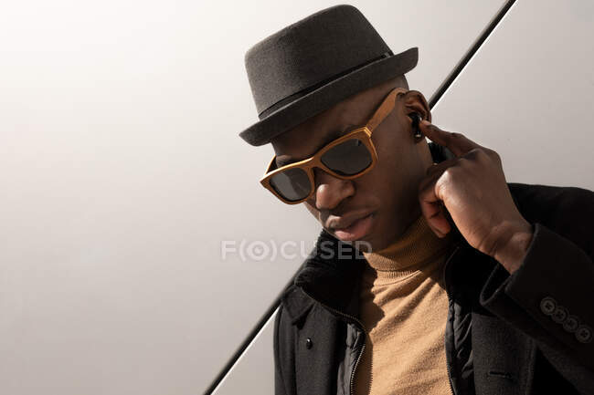 Trendy confident African American male in hat and sunglasses standing against gray wall and looking away — Stock Photo