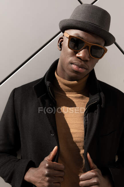 Moda confiante afro-americano masculino em chapéu e óculos de sol em pé contra a parede cinza e olhando para a câmera — Fotografia de Stock