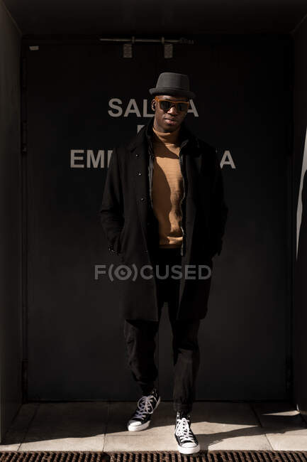 Full body trendy serious African American male in hat and sunglasses standing with hands in pockets on sunny street and looking at camera — Stock Photo