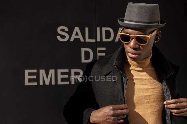Trendy confident African American male in hat and sunglasses standing against gray wall and looking away — Stock Photo