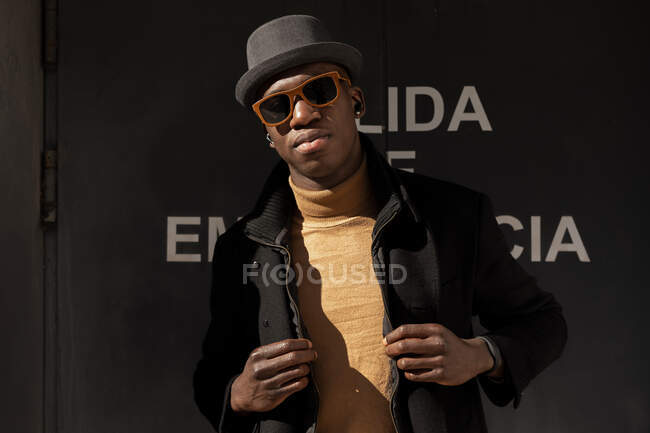Впевнений афроамериканець у капелюсі і сонцезахисних окулярах стоїть проти сірої стіни і дивиться на камеру — стокове фото