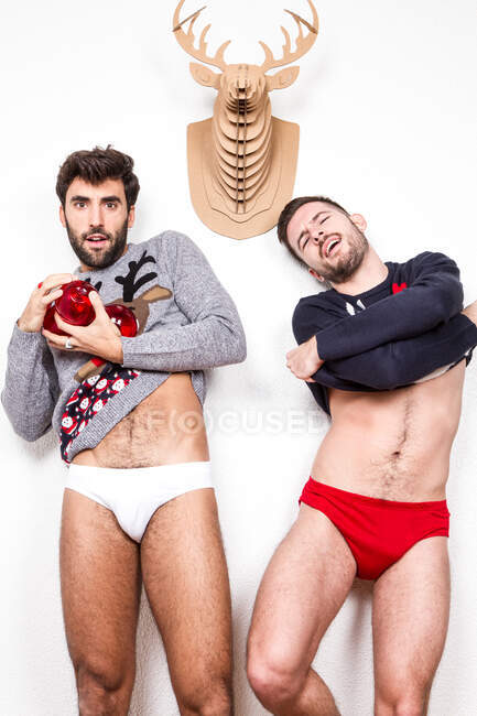 Casal de adultos amantes gays em roupas íntimas e camisolas Xmas com bolas vermelhas decorativas em pé contra a parede branca com cabeça de veado artificial — Fotografia de Stock