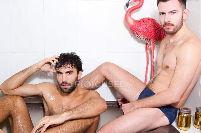 Багаторасові бородаті гей-коханці з голими торнами відпочивають разом у ванні з рожевим фламінго — стокове фото