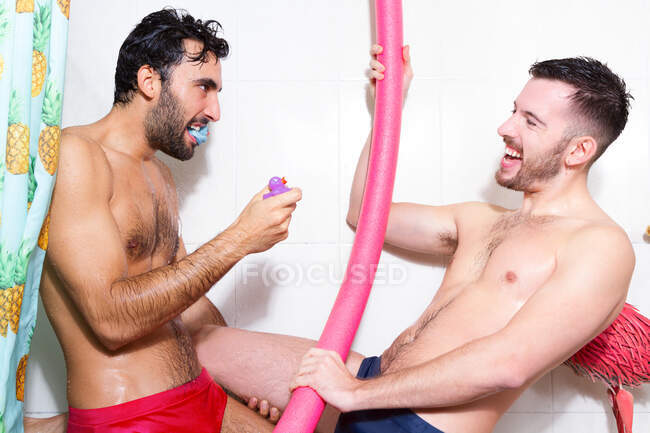 Вид збоку веселих різноманітних бородатих гомосексуальних хлопців з голими торсами, які розважаються з гумовими іграшками та локшиною для басейну, приймаючи душ разом у ванній — стокове фото