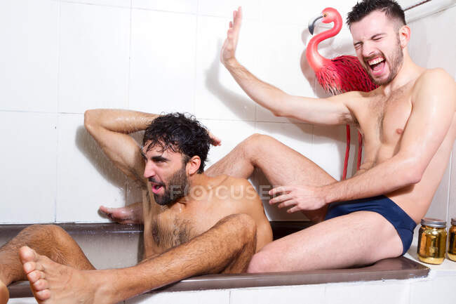 Vista laterale di adulti barbuti senza maglietta fidanzati omosessuali multirazziali che si fanno la doccia seduti nella vasca da bagno e si divertono insieme — Foto stock