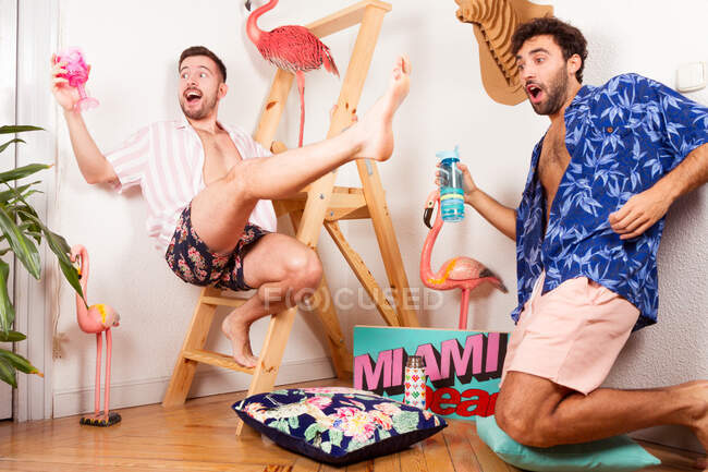 Divertido emocionado diversos adultos homosexuales novios en trajes de verano con bebidas fingiendo estar en la playa con flamenco rosa y divertirse juntos - foto de stock