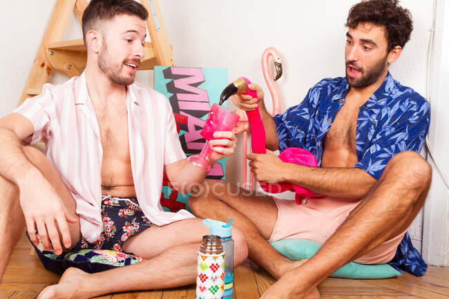 Молодой гей наливает напиток взволнованному парню, притворяясь, что веселится на летнем тропическом пляже вместе — стоковое фото