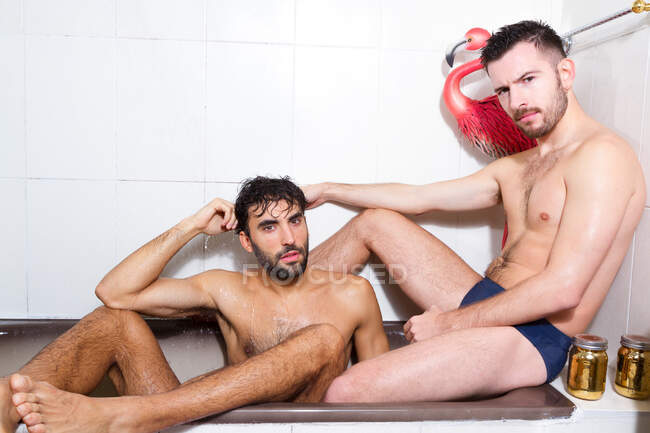 Багаторасові бородаті гей-коханці з голими торнами відпочивають разом у ванні з рожевим фламінго — стокове фото