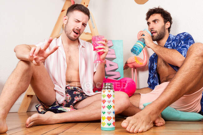 Funny excited diverse adult homosexual boyfriends in summer outfits with drinks pretending being on beach with pink flamingo and having fun together — Stock Photo