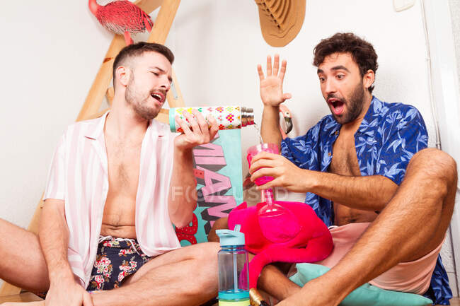 Молодий гей-чоловік виливає напій збудженому хлопцеві, вдаючи, що весело провести час на літньому тропічному пляжі разом — стокове фото