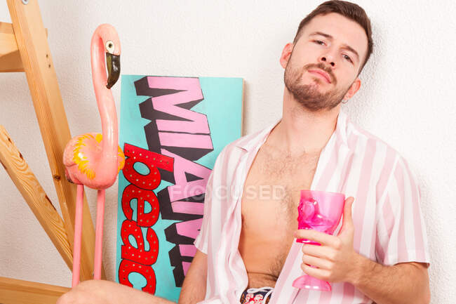 Jovem barbudo homossexual cara em roupa de verão beber coquetel de cálice rosa enquanto sentado no chão em casa e fingindo estar na praia — Fotografia de Stock