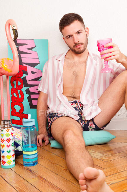 Joven barbudo homosexual en traje de verano bebiendo cóctel de copa rosa mientras está sentado en el suelo en casa y fingiendo estar en la playa - foto de stock