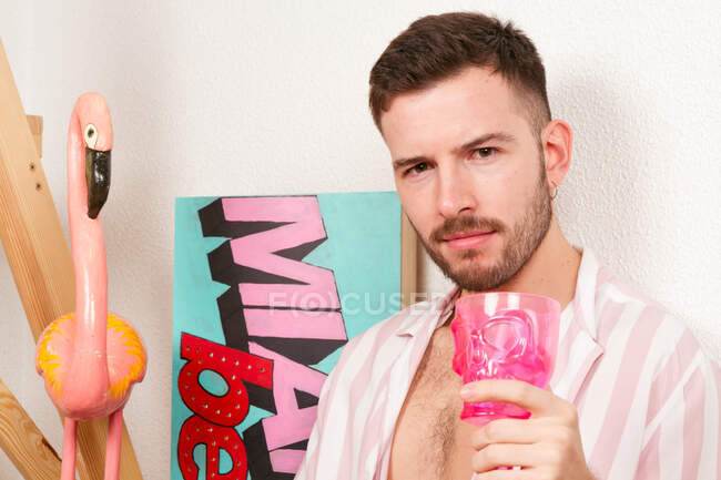 Junger bärtiger homosexueller Kerl im Sommer-Outfit trinkt Cocktail aus rosa Becher, während er zu Hause auf dem Boden sitzt und vorgibt, am Strand zu sein — Stockfoto