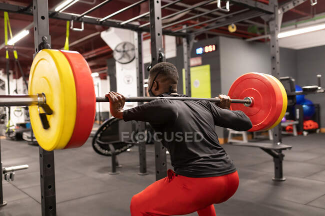 Atrás ver a un joven atleta afroamericano en ropa deportiva y mascarilla levantando pesadas barras de metal mientras entrenaba en el gimnasio durante la pandemia de coronavirus - foto de stock