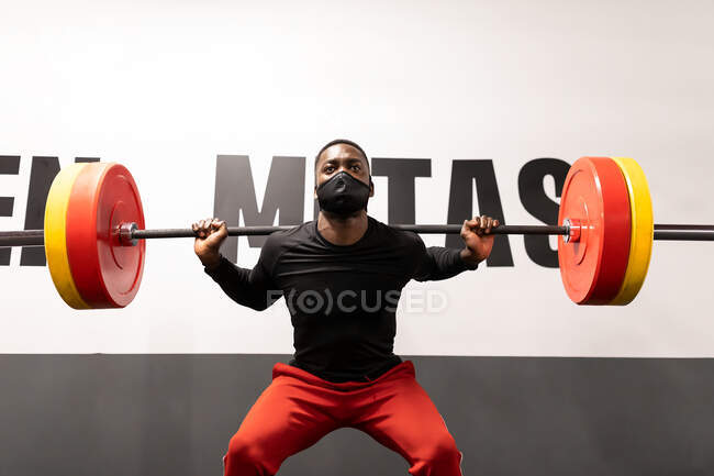 Determined young African American male athlete in activewear and face mask lifting heavy metal barbell while training in gym during coronavirus pandemic — Stock Photo