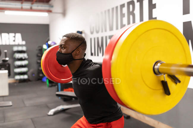 Side view young African American male athlete in activewear and face mask lifting heavy metal barbell while training in gym during coronavirus pandemic — Stock Photo