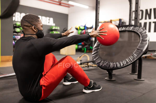 Вид сбоку на молодого черного спортсмена в спортивной и медицинской маске, делающего упражнения на прессе с мячом во время функциональной тренировки в современном тренажерном зале — стоковое фото