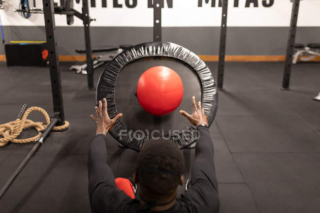 Vista posterior de un joven deportista negro en ropa deportiva haciendo ejercicios abdominales con balón de medicina durante el entrenamiento funcional en el gimnasio moderno - foto de stock