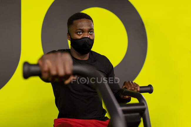 Сконцентрований молодий афроамериканець спортсмен в спортивній манері і маска на велосипедній машині під час тренування в спортзалі на яскраво-жовтому фоні — стокове фото