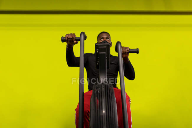 Low angle of concentrated young African American male athlete in sportswear and face mask exercising on cycling machine during training in gym against bright yellow background — Stock Photo