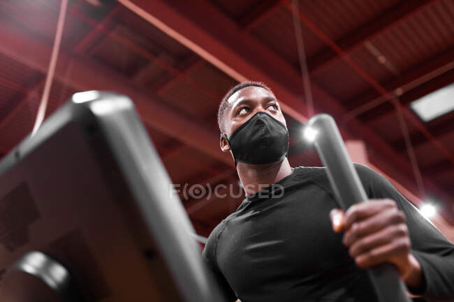 Снизу афроамериканский спортсмен в маске делает кардиотренировку на эллиптической машине в современном тренажерном зале — стоковое фото