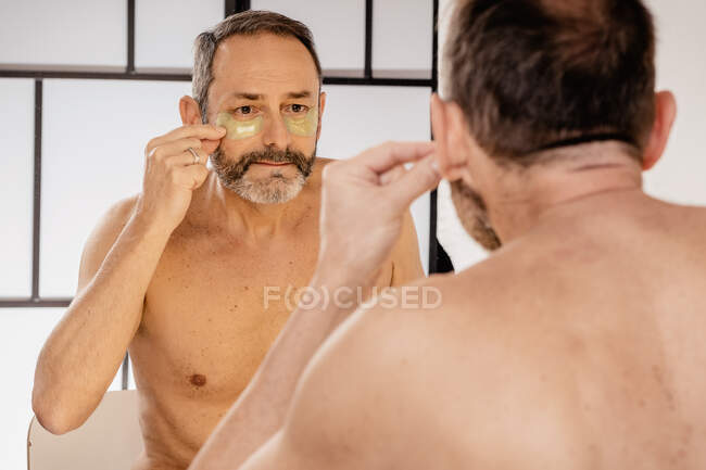 Бородатый мужчина среднего возраста с обнаженным туловищем, надевающий повязку на кожу, глядя в зеркало дома — стоковое фото