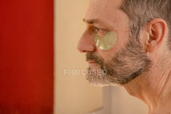 Боковой вид обрезанного бородатого мужчины средних лет с обнаженным туловищем, накладывающим повязку на кожу, глядя в зеркало дома — стоковое фото