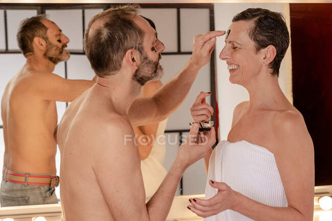 Sid-Ansicht von fröhlichen Mann in Handtuch Anwendung schwarzer abziehen Maske auf das Gesicht der weiblichen Geliebten, während sie einander im Spiegel reflektiert zu Hause — Stockfoto