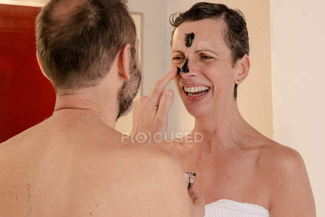 Vista de Sid de alegre macho en toalla aplicando la máscara de piel negra en la cara de la mujer amada mientras se miran reflejándose en el espejo en casa - foto de stock