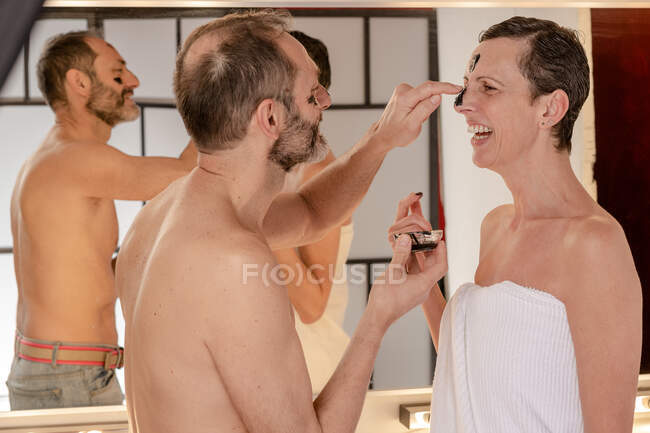 Sid view of cheerful male in towel applying black peel off mask on face of female beloved while looking at each other reflecting in mirror at home — Stock Photo