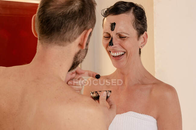 Sid view of cheerful male in towel applying black peel off mask on face of female beloved while looking at each other reflecting in mirror at home — Stock Photo