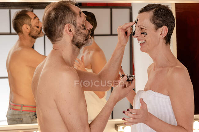 Sid-Ansicht von fröhlichen Mann in Handtuch Anwendung schwarzer abziehen Maske auf das Gesicht der weiblichen Geliebten, während sie einander im Spiegel reflektiert zu Hause — Stockfoto