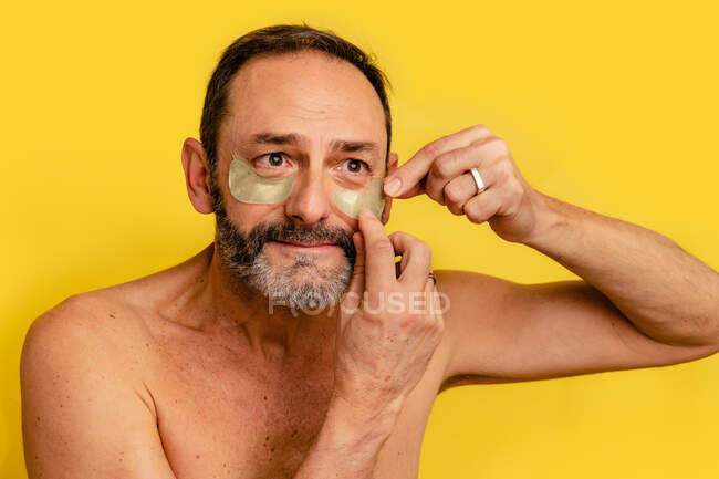 Чоловік середнього віку з голим торсом наносить плями очей на шкіру, дивлячись на жовтий фон — стокове фото