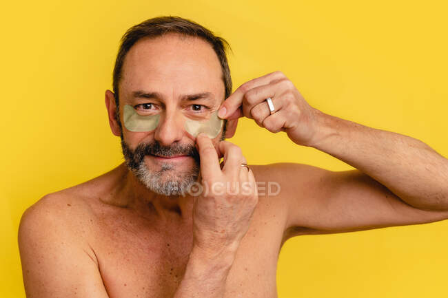 Middle aged male with naked torso applying eye patches on skin while looking at camera on yellow background — Stock Photo