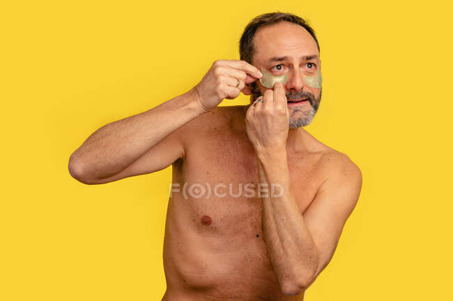 Middle aged male with naked torso applying eye patches on skin while looking away on yellow background — Stock Photo