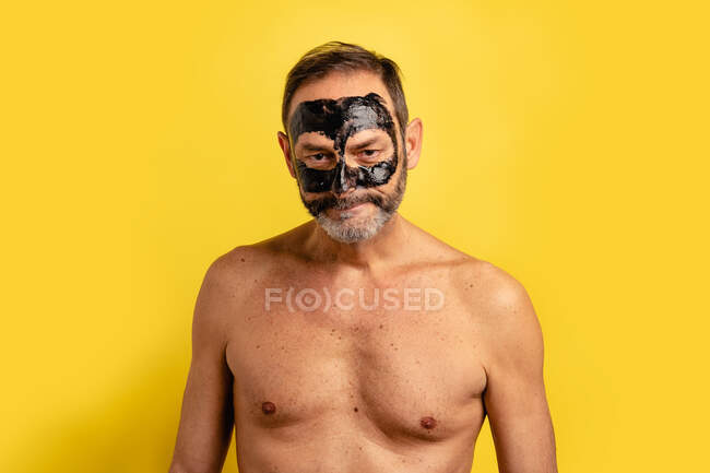 Macho de meia idade mostrando preto descascar máscara no rosto enquanto olha para a câmera no fundo amarelo — Fotografia de Stock