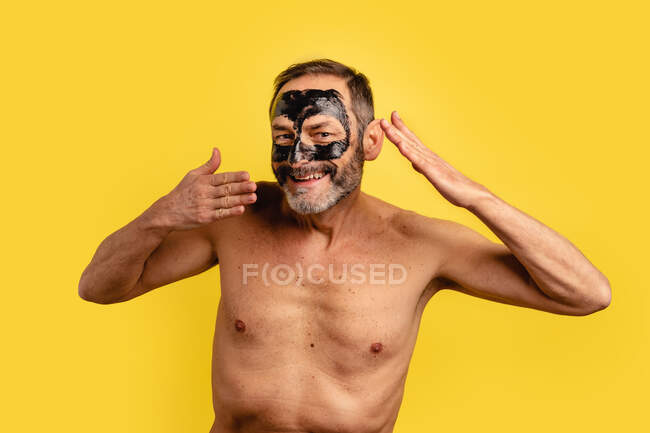 Sorrindo macho de meia-idade mostrando preto descascar máscara no rosto enquanto olha para a câmera no fundo amarelo — Fotografia de Stock