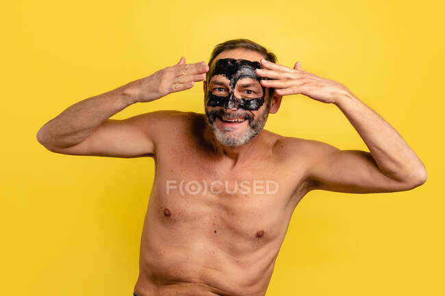 Мужчина средних лет улыбается, показывая черную кожуру с маски на лице, глядя на камеру на желтом фоне. — стоковое фото