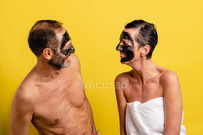 Smiling middle aged couple in towel with peel off mask on faces standing looking at each other while on yellow studio background — Stock Photo