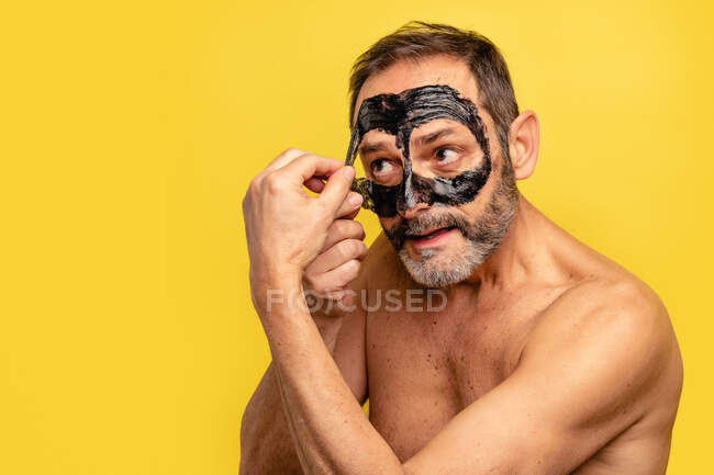 Varón de mediana edad con torso desnudo pelando la máscara negra de la cara mientras mira hacia otro lado en el fondo amarillo - foto de stock