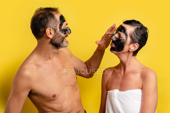 Macho alegre em toalha aplicando a máscara de casca preta no rosto da amada feminina enquanto olham um para o outro no fundo amarelo — Fotografia de Stock