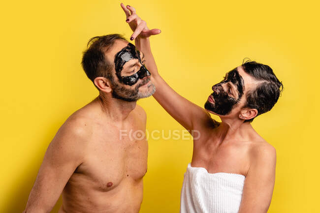 Mulher alegre em toalha aplicando a máscara de casca preta no rosto do macho amado enquanto olha um para o outro no fundo amarelo — Fotografia de Stock