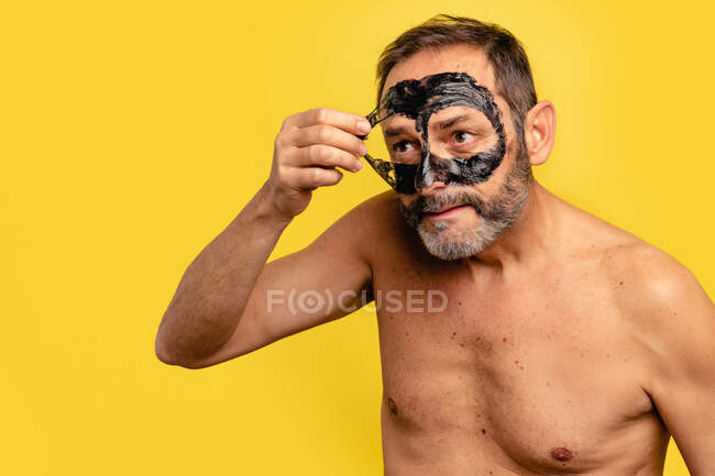 Middle aged male with naked torso peeling black mask off face while looking away on yellow background — Stock Photo