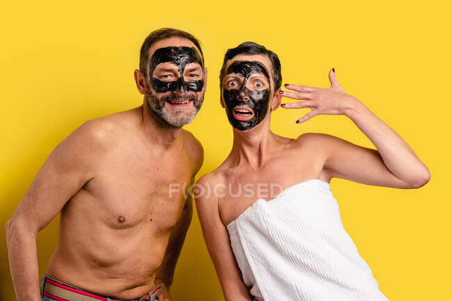 Усміхнений чоловік з голим торсом біля здивованої дівчини в рушнику, що показує відшаровування маски на обличчі, дивлячись на камеру на жовтому фоні — стокове фото