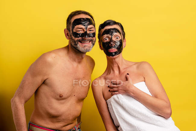 Улыбающийся мужчина с обнаженным туловищем рядом с изумленной девушкой в полотенце, показывающий снятую маску на лице, глядя на камеру на желтом фоне — стоковое фото